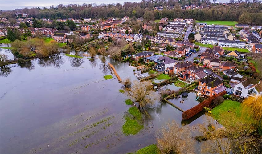 A birds eye view image taken from height overlooking houses which have been flooded. In front of the houses is a large field which is flooded with water. 