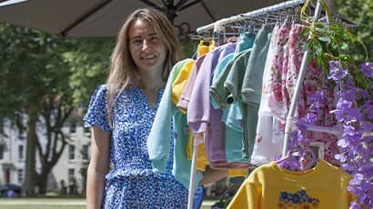 Photo shows a lady smiling in front of a rack of clothes she's made