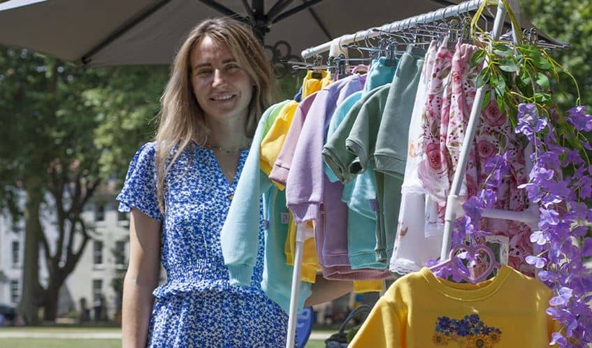 Photo shows a lady smiling in front of a rack of clothes she's made