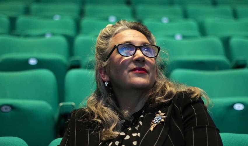 A woman with glasses in a theatre sitting amongst a bank of green seats. 