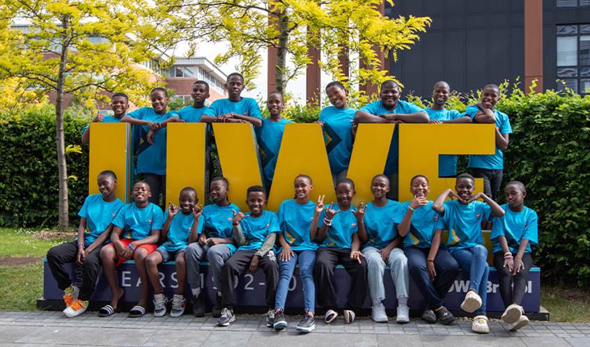 A group of children wearing bright blue tshirts sit outside on a giant UWE logo, smiling at the camera.