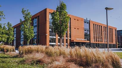 A landscape shot taken outside showing trees and shubbery in the foreground and a large orange building in the background.