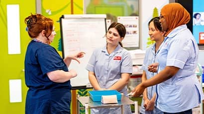 Student healthcare professionals in uniform stand listening to a lecturer in blue scrubs. 