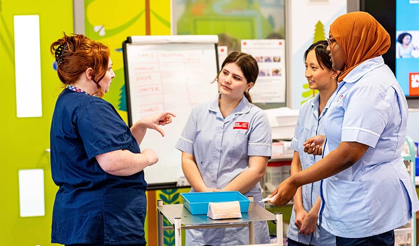 Student healthcare professionals in uniform stand listening to a lecturer in blue scrubs.