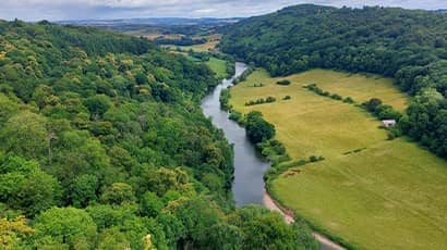 A wide landscape shot taken from a high vista overlooking a river running between green fields and woodlands.