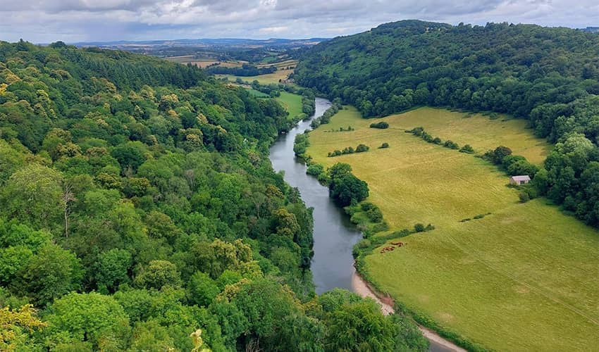 A wide landscape shot taken from a high vista overlooking a river running between green fields and woodlands. 