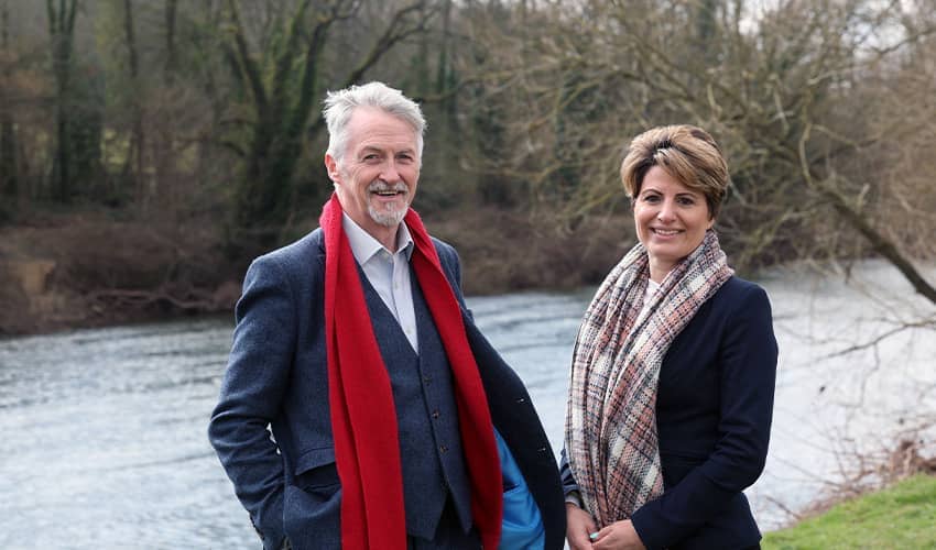 A man and a woman in suit blazers and scarves smile at the camera. They are stood outside next to a large river with woodlands behind it. 