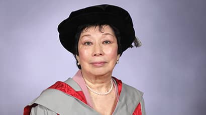 A woman wearing university graduation gowns and a hat smiles at the camera with a grey background behind. 
