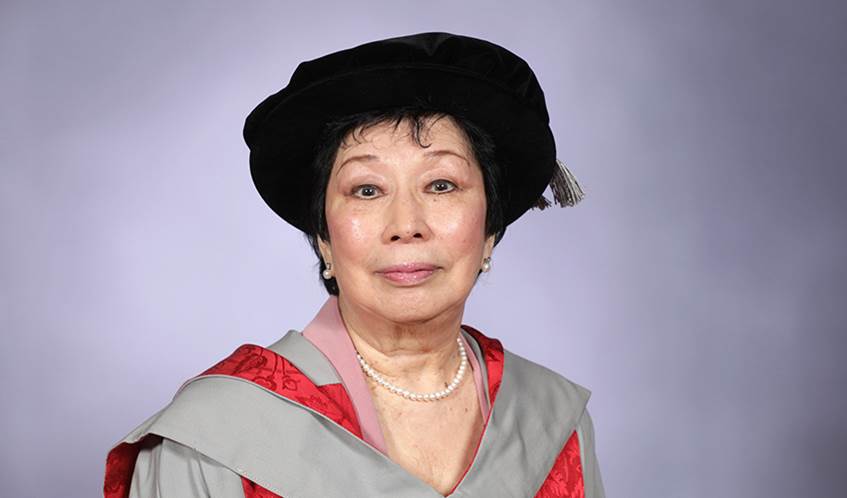A woman wearing university graduation gowns and a hat smiles at the camera with a grey background behind.