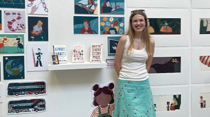 A woman wearing a white top and turquoise skirt is photographed with her illustration designs on a wall behind her.