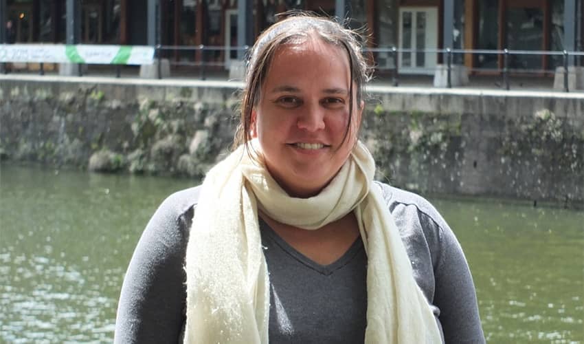  A person wearing a light scarf and dark top stands outdoors by a river or canal, with a building featuring large windows and bunting visible across the water in the background.
