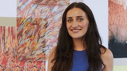 A woman with dark hair wearing a blue top smiles at the camera, with artwork behind her