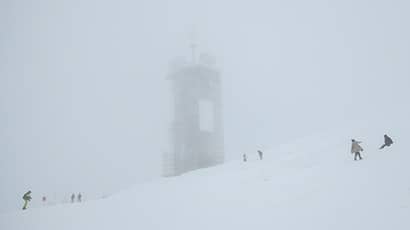 People on a snow-covered slope in the fog with a tall tower in the background.