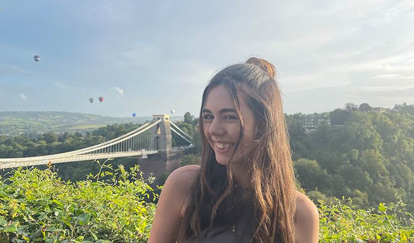 UWE Bristol student Emily Annakin stands smiling with the Clifton Suspension Bridge behind her with hot air balloons flying in the distance