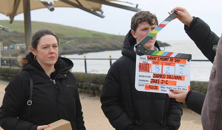 Two actors stand behind a clapperboard during outdoor filming by the coast.