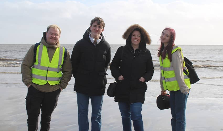 Four people standing on a beach shoreline, two wearing high‑visibility vests and one holding filming equipment.