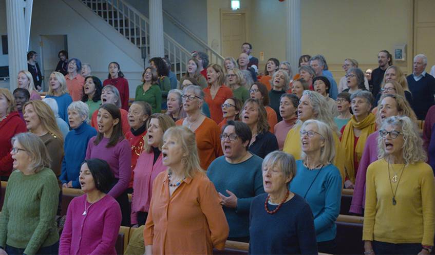 A group of people are enthusiastically singing, while wearing brightly coloured jumpers.