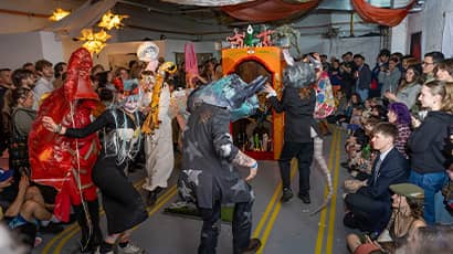 A crowd watches a group of students artistically dressed as they dance around a studio. 