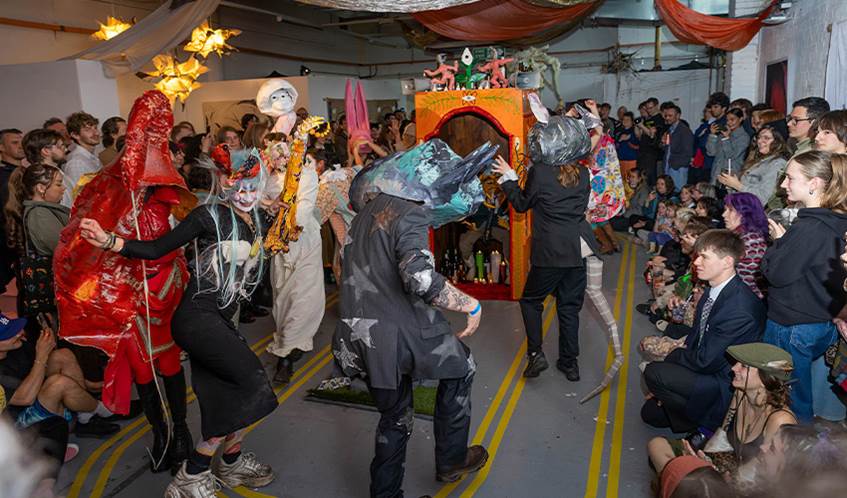 A crowd watches a group of students artistically dressed as they dance around a studio.