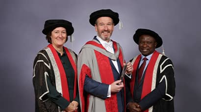 Three people stand together smiling at the camera wearing graduation robes. 