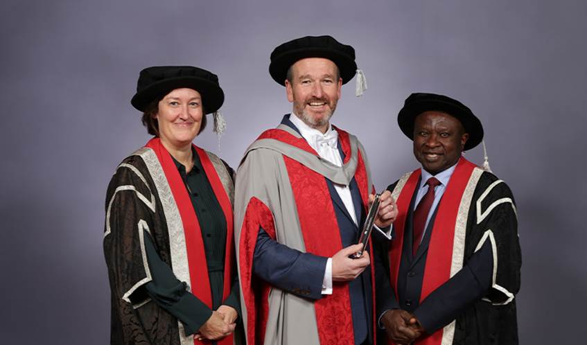 Three people stand together smiling at the camera wearing graduation robes.