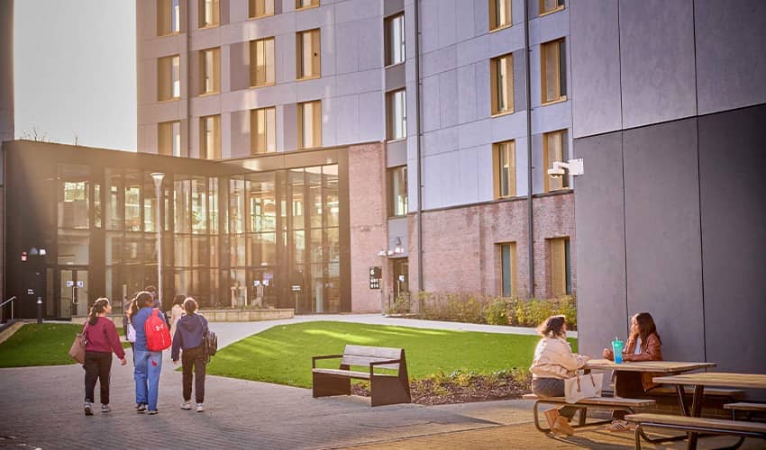 A wide shot showing a group of young people walking towards a large building with big glass windows on a university campus. 