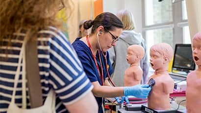 A medical clinician demostrates uses a stethescope on a child manequin while a young adult watches on.