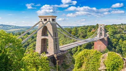 The image shows the Clifton Suspension Bridge, a famous suspension bridge spanning the Avon Gorge and the River Avon in Bristol, England. The bridge features two large stone towers with cables extending from them to support the roadway. The surrounding area is lush with greenery and trees, and there are hills in the background under a partly cloudy sky.