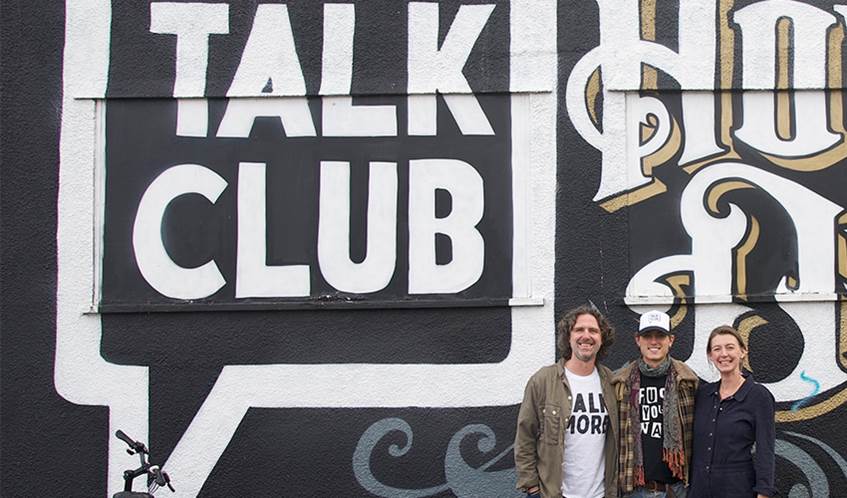 Three people stood in front of a black wall which has a speech bubble and the words Talk Club painted on it