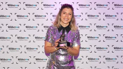 A woman wearing a shiny dress smiles at the camera, while holding an award