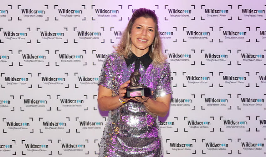 A woman wearing a shiny dress smiles at the camera, while holding an award