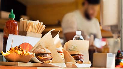An outdoor food stall with two burgers and a bowl of chips lined up next to ketchup and napkins, with a person blurred in the background working on the stall. 