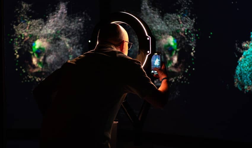 Person photographing an illuminated circular display with a smartphone in a dark immersive exhibition.