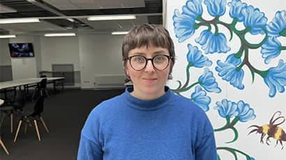 Person in a classroom smiling at the camera. The person is wearing a blue jumper and glasses and is stood in front of a white wall that has blue flower and bumblebee decorations. 