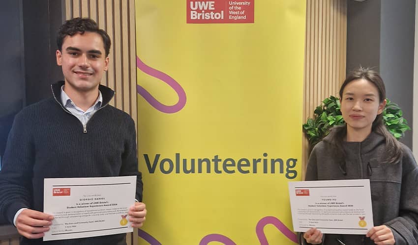 Two people stand in front of a UWE Volunteering banner. They are both holding certificates. One is wearing a grey jumper and light blue shirt and the other is wearing a dark grey coat. 