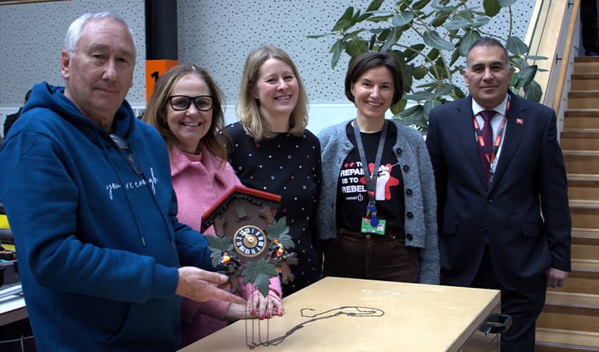 Five peple stood in a row smiling at the camera, with one man at the front of the group holding a cuckoo clock.