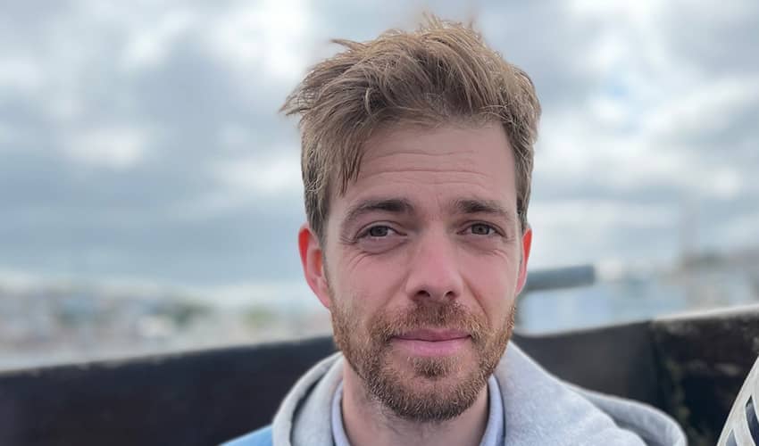 A man with light brown hair stands outdoors, wearing a light grey hoodie.