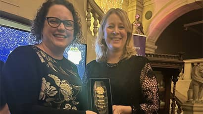 Two women stand holding an award between them smiling at the camera. They are both wearing smart black dresses. 