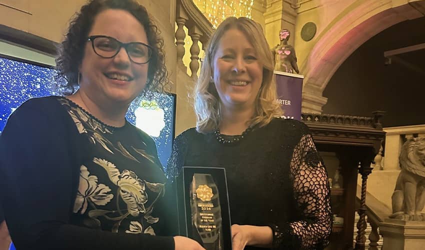 Two women stand holding an award between them smiling at the camera. They are both wearing smart black dresses. 