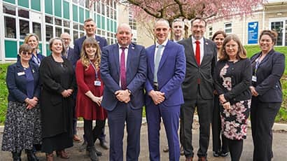 A group of 13 people wearing smart suits or work attire stood outside a school setting underneath a large blossom tree. 