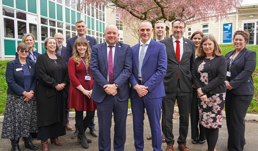 A group of 13 people wearing smart suits or work attire stood outside a school setting underneath a large blossom tree. 