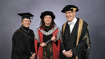 Two women and a man wearing gowns and mortar boards looking at the camera against a grey background