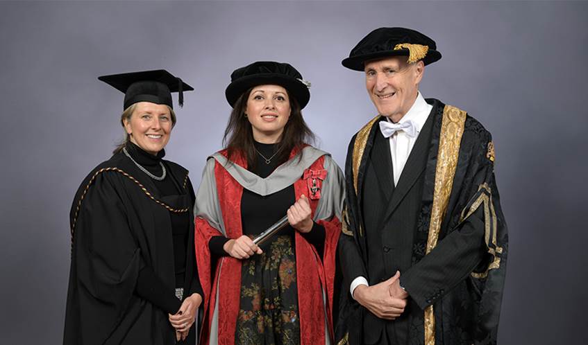 Two women and a man wearing gowns and mortar boards looking at the camera against a grey background