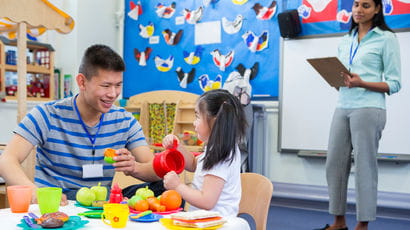 Male teacher playing with a little girl. Both are seated at a child's table. There is a female teacher in the background with a clipboard who is watching them.