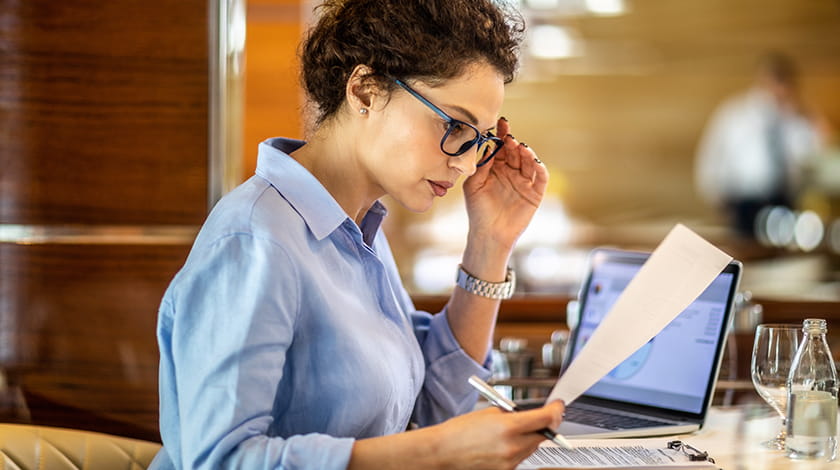 Female entrepreneur reading a paper document while sitting down. She is holding her reading glasses. A laptop, glass and water bottle are on the table.