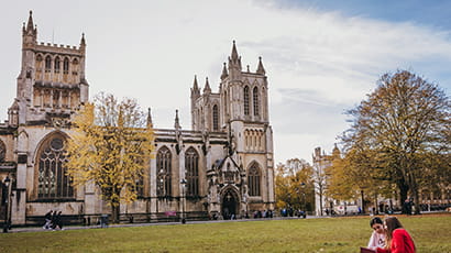 Two females sitting on the lawn in front of Bristol Cathedral against a blue sky.