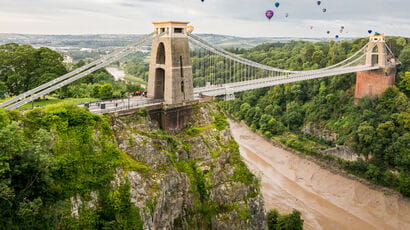 Aerial shot of the Clifton Suspension Bridge over Avon Gorge with hot air balloons in the background.