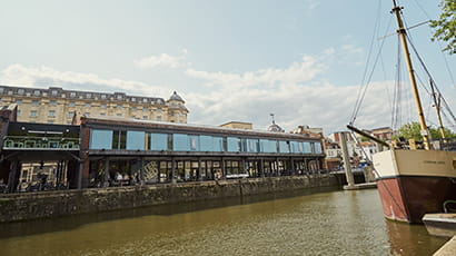 Wide angle view of Watershed in the daylight at Bristol Harbourside with a balcony in the background and a ship in the foreground.