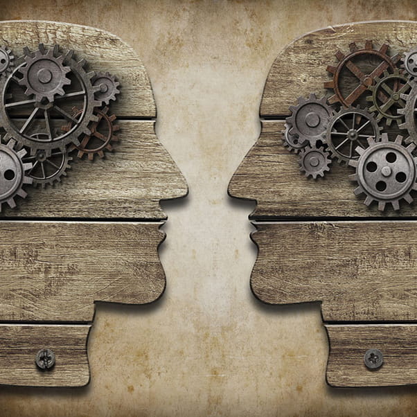 Two wooden heads in profile facing each other with multiple cog wheels representing their brains.