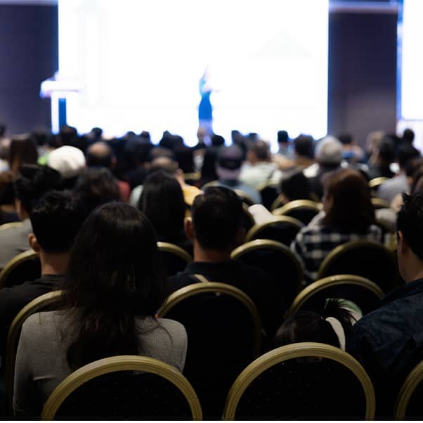 Rear view show of delegates in a conference hall with a speaker on stage.